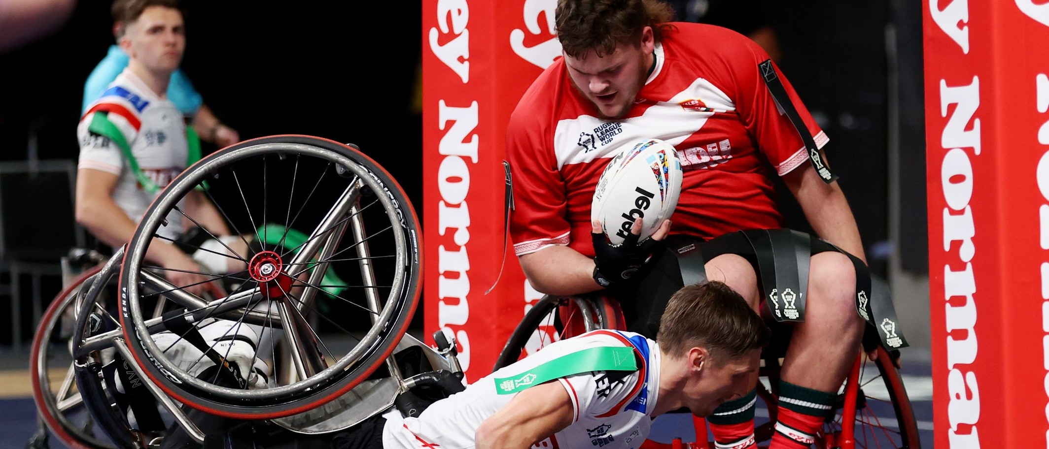A Wales wheelchair rugby league player in a red shirt holds the ball over the line as an England player falls in front of him