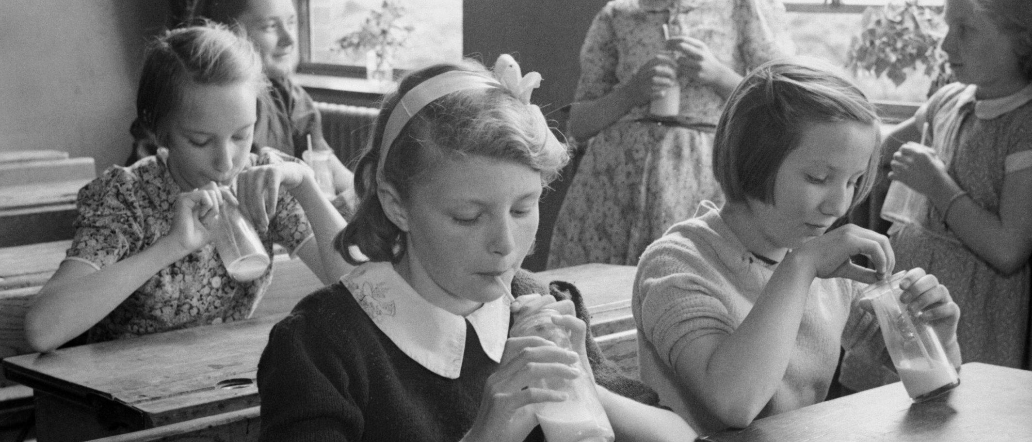 Girls drink milk through straws from small glass bottles at wooden school desks. Archive picture from 1944 in Hertfordshire, not a Welsh school. Photo: Ministry of Information Photo Division, Imperial War Museums D 20552, public domain, via Wikimedia Commons