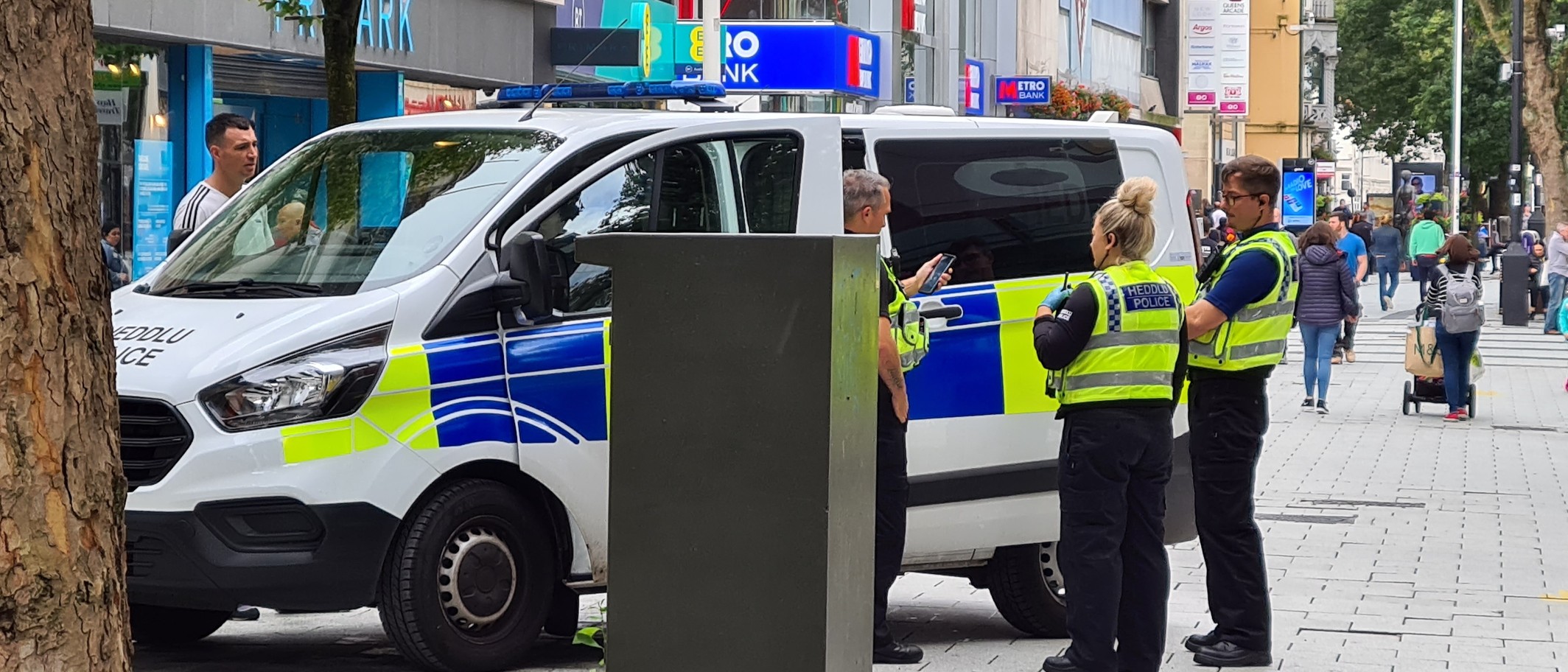 Three South Wales Police officers beside a marked police van in Cardiff city centre. Photo: Scglossop1, CC BY-SA 4.0, via Wikimedia Commons.