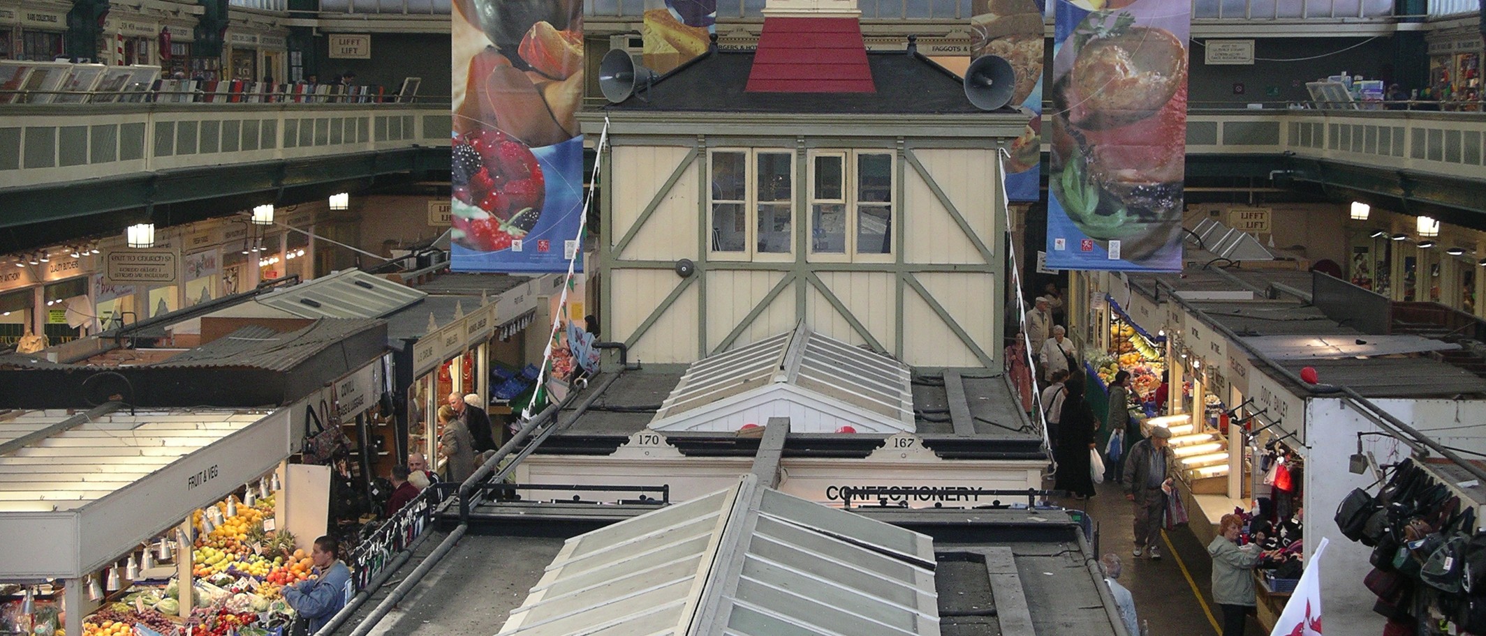 Archive interior of Cardiff Market on 7 November 2005, with food stalls and hanging produce banners. Velela / Wikimedia Commons, public domain; cropped.